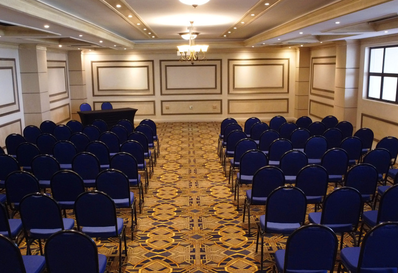 Conference room with rows of blue banquet chairs, patterned carpet, paneled walls and a front speaker table under chandelier.