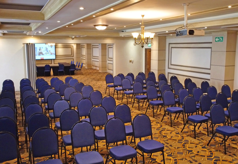 Hotel meeting room set theater-style with rows of blue chairs, ceiling projector and screen, carpet and decorative wall panels.