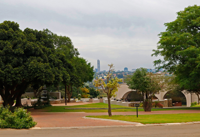 Landscaped hotel grounds with manicured lawns, shaded trees and fountain, arched terrace overlooking a distant city skyline.