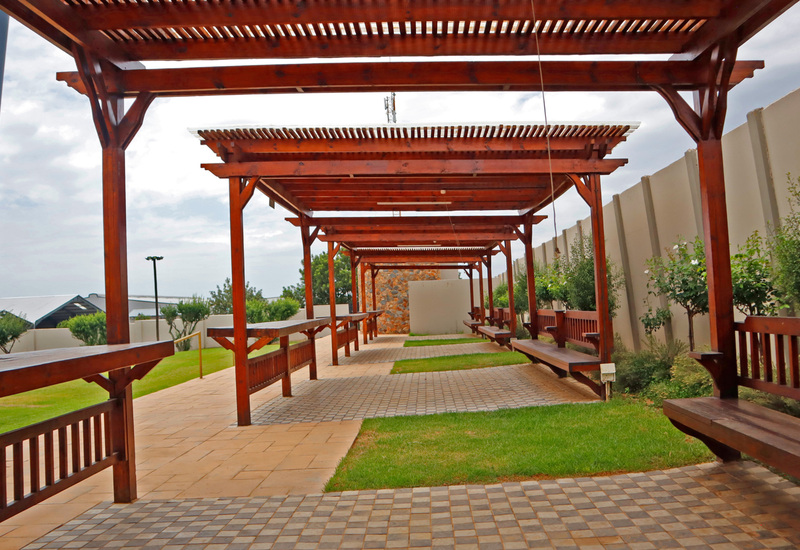 Shaded outdoor pergola with teak benches and bar tables along a tiled walkway, grassy patches and landscaped privacy wall.