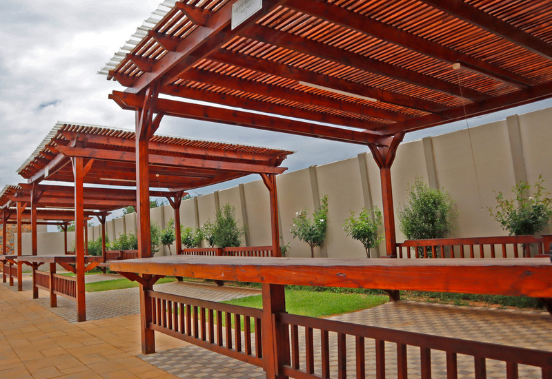 Wooden pergola-covered outdoor dining area with long communal tables and benches, tiled paths, small landscaped trees.