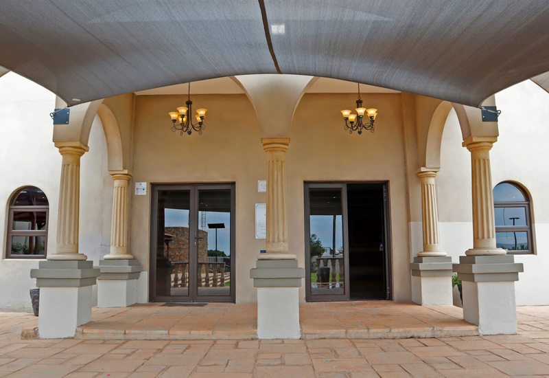 Mediterranean-style hotel entrance with arched portico, fluted columns, hanging chandeliers, shaded canopy and double glass doors opening to lobby.