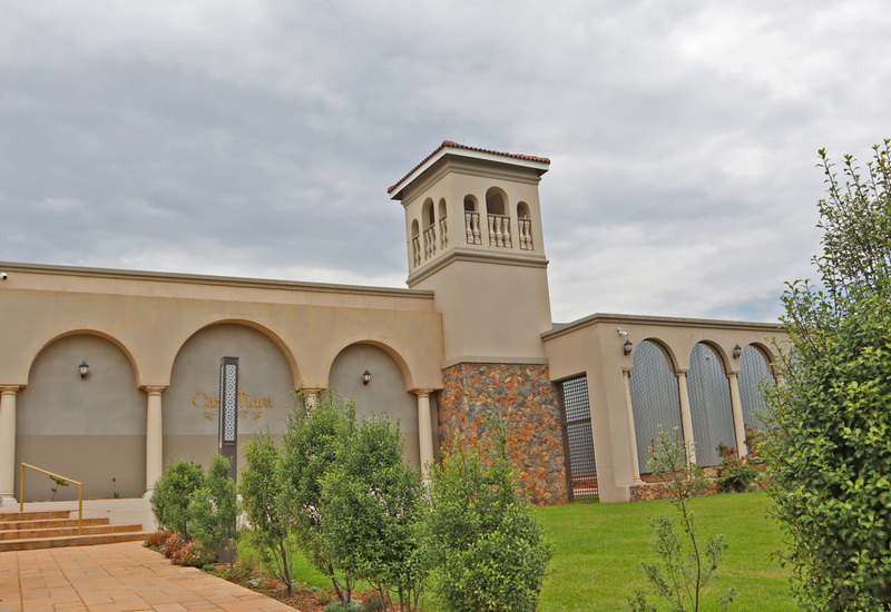 Mediterranean hotel facade with arched colonnade, tiled tower, stone base and lanterns; steps from a tree-lined paved walkway.