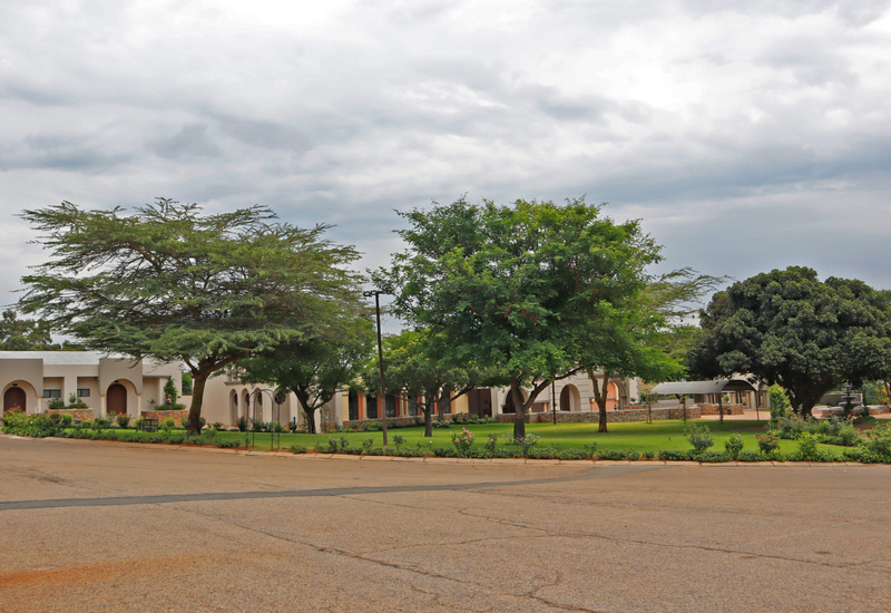 Low-rise hotel with arched entrances around a manicured lawn courtyard, shaded by large trees, garden beds, covered drop-off and fountain.