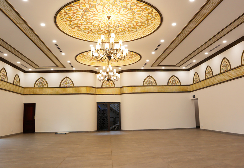 Ornate hotel banquet hall with high ceilings, gold patterned ceiling medallions, triple chandeliers, arched windows and wide event-ready floor.