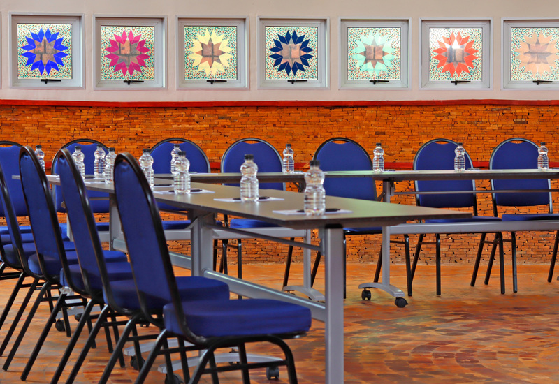 Conference room with blue banquet chairs and long tables on casters, bottled water at each seat, stained-glass windows and brick wall.