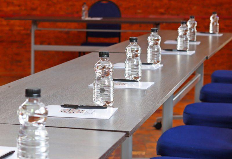 Conference table set for meeting with bottled water, pens and notepads, blue padded chairs and exposed brick wall backdrop