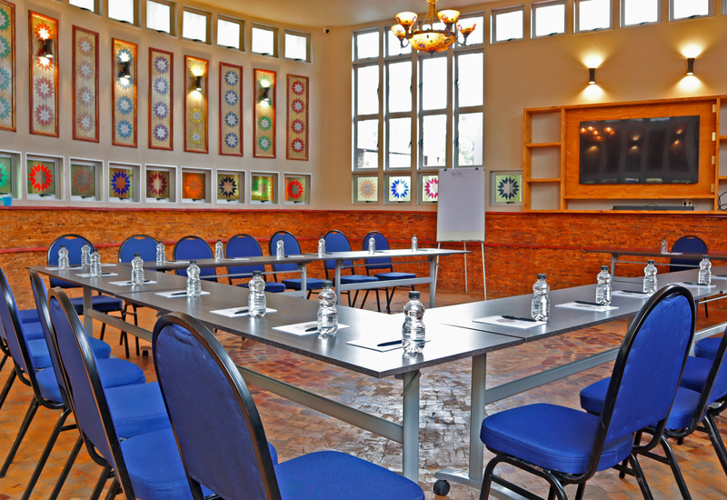 U-shaped meeting room with blue chairs, bottled water and notepads, wall TV, flipchart, tall windows and decorative panels.