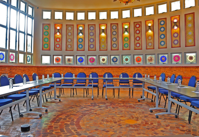 Bright circular meeting room with U-shaped tables, blue chairs, water bottles, mosaic floor and star stained-glass windows
