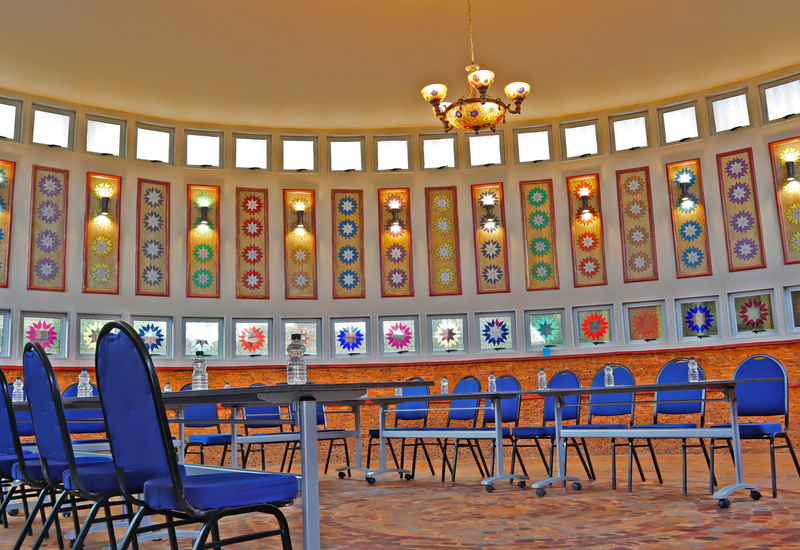 Circular hotel meeting room with rows of blue chairs and tables, decorative stained-glass star panels, warm chandelier light.