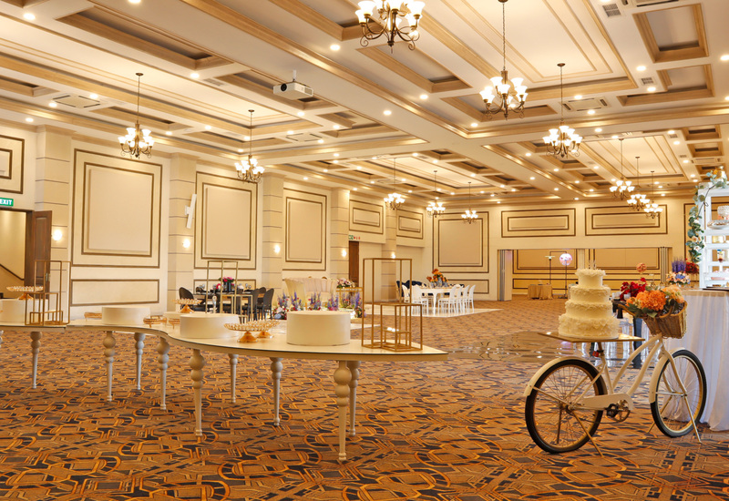 Grand ballroom with coffered ceiling, chandeliers, dessert tables, floral displays and tiered wedding cake on a bicycle.