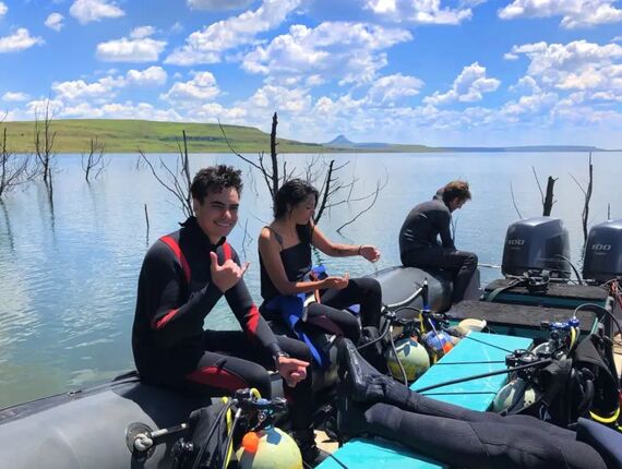 Guests in wetsuits on a dive boat with scuba tanks, preparing for a lake dive amid calm water, dead trees and rolling hills.