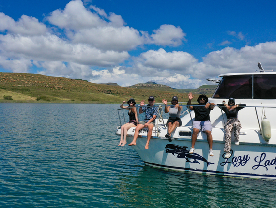 Guests waving from the bow of Lazy Lady IV yacht, legs dangling over turquoise lake under a sunny blue sky