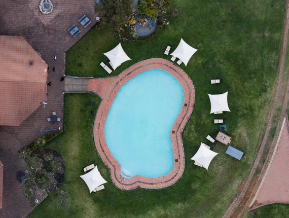Curved outdoor pool with brick surround, white canopy shade tents and loungers on a green lawn by terracotta-roofed hotel.