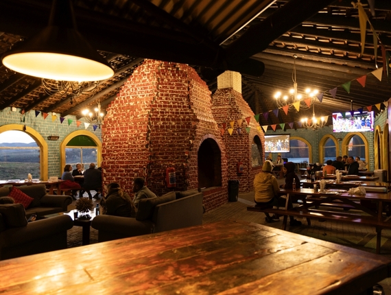 Rustic lounge-restaurant with large red-brick fireplace, wooden beams, communal tables and sofas, guests dining and chatting by arched windows.