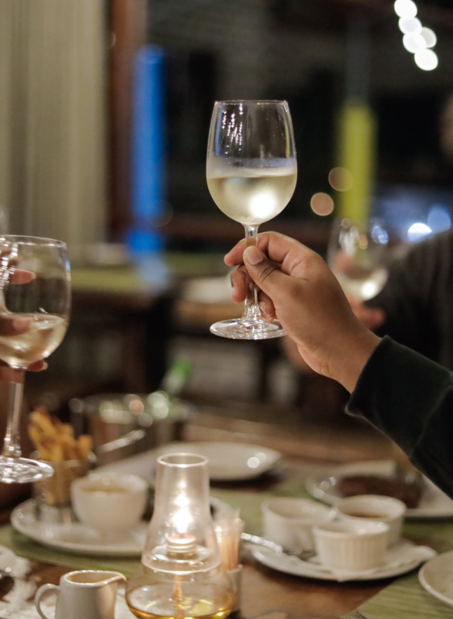 Guests toasting with chilled white wine over a candlelit dining table with shared plates, fries and warm string lights.