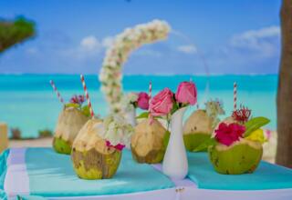 Fresh coconuts with flowers and striped straws on a beachfront table with turquoise ocean and floral arch in the background