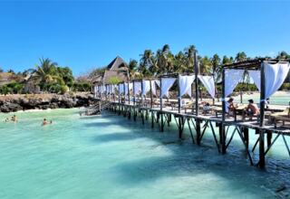 Wooden pier with shaded cabanas and lounge chairs over turquoise water, guests relaxing and swimming by rocky beachfront with palm trees