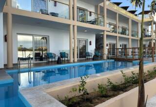 Modern hotel with outdoor pool, balcony seating, glass railings, and patio chairs overlooking tropical palms and beach view
