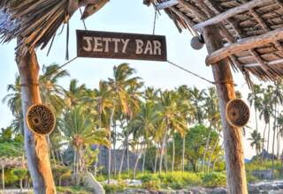 Rustic wooden Jetty Bar sign hanging under a thatched roof with palm trees and tropical greenery in the background