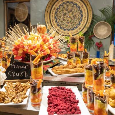 Colorful buffet with fruit skewers, peanut crust treats, assorted pastries, and a smiling chef in warm, decorative setting