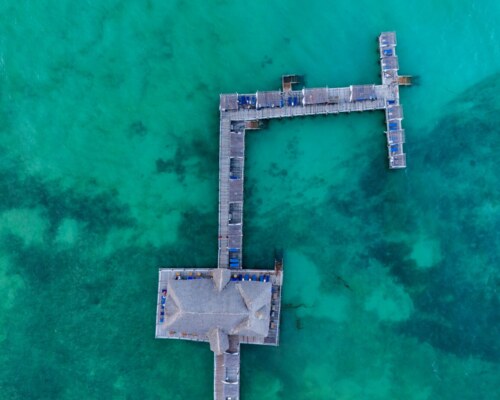 Aerial view of a wooden pier with lounge chairs extending over clear turquoise ocean water, perfect for relaxation and sunbathing