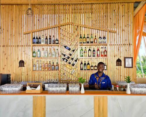 Bright hotel bar with wooden slat wall featuring assorted liquor bottles, glassware, green plants, and a smiling bartender in blue attire