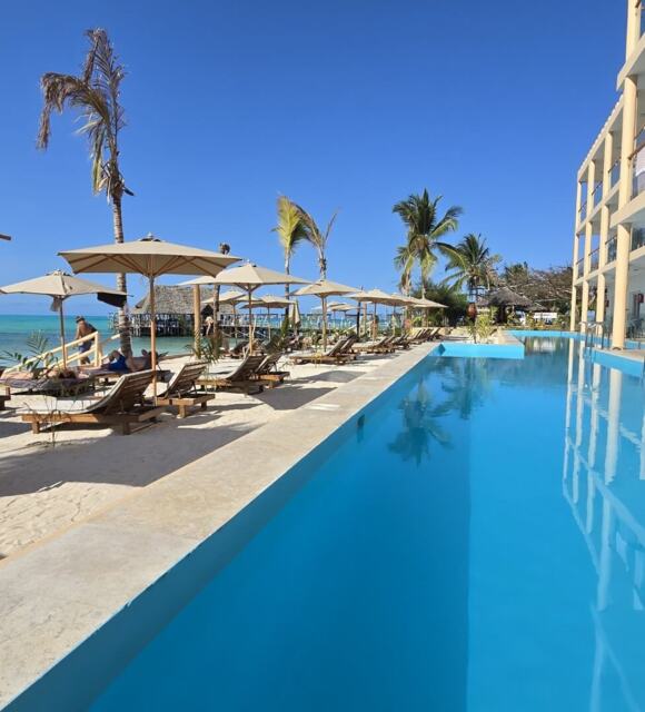 Beachfront pool with lounge chairs and umbrellas on sandy shore, adjacent to a modern hotel with balconies and ocean views