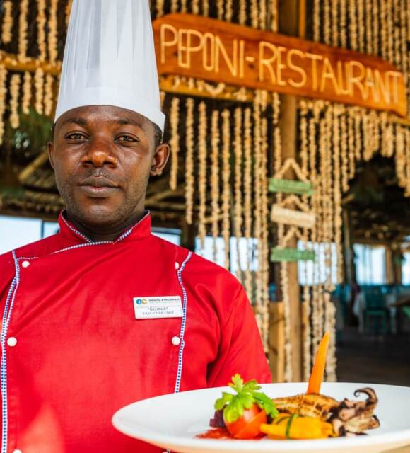 Executive chef in red uniform presenting a gourmet seafood dish at rustic Peponi Restaurant with natural decor and open-air seating
