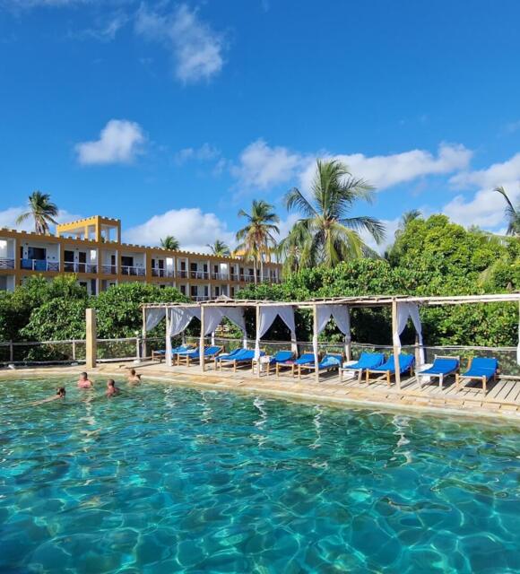 Outdoor swimming pool with guests enjoying the water, shaded lounge chairs with white canopies, surrounded by tropical palm trees and a three-story hotel building
