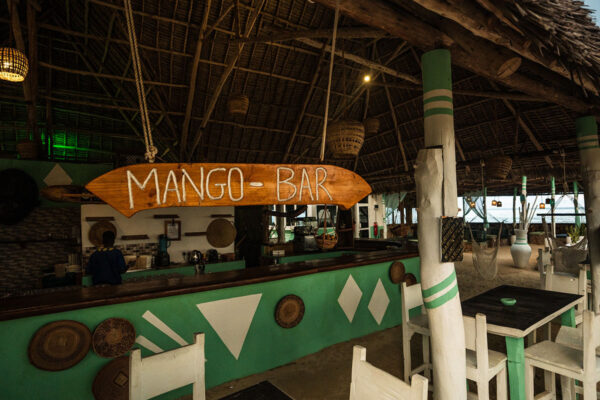 Rustic open-air Mango Bar with thatched roof, green and white decor, wooden tables and chairs, and a relaxed tropical ambiance
