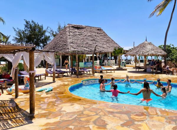 Guests enjoy a group water exercise in a sunny round pool surrounded by thatched-roof cabanas and lounge chairs near the beach