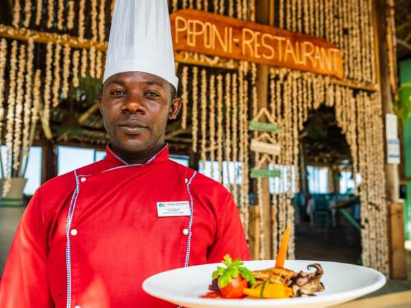 Executive chef in red uniform presenting a gourmet seafood dish at rustic Peponi Restaurant with natural decor and open-air seating