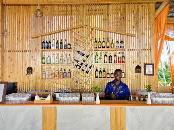 Bright hotel bar with wooden slat wall featuring assorted liquor bottles, glassware, green plants, and a smiling bartender in blue attire