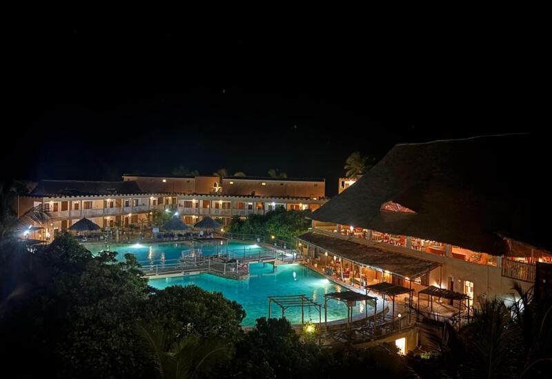 Night view of a tropical resort featuring a large illuminated pool surrounded by rooms with balconies and a thatched-roof restaurant area