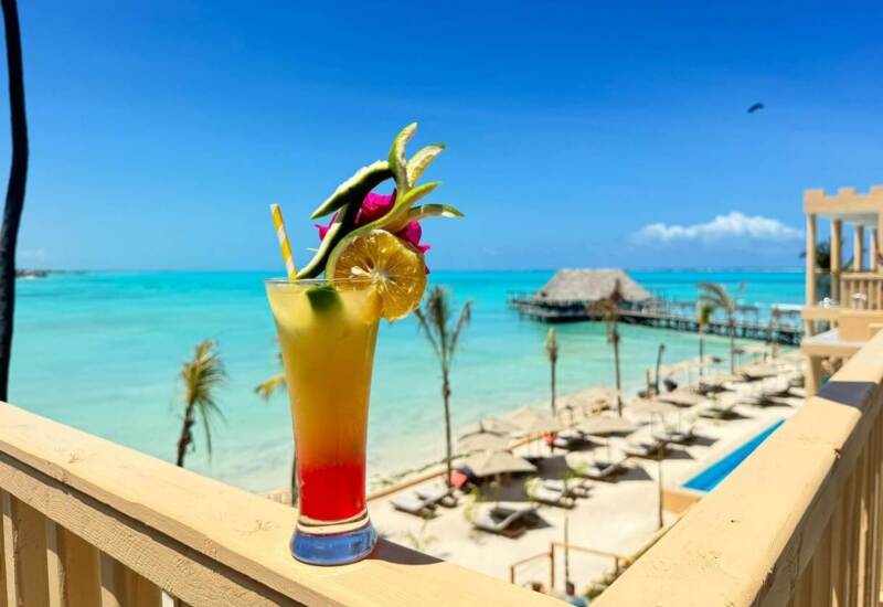 Tropical cocktail garnished with lemon and dragon fruit on balcony railing overlooking beachfront resort with lounge chairs and turquoise ocean
