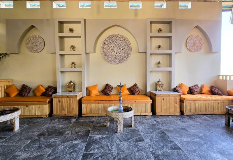 Cozy lounge with wooden bench seating, orange cushions, decorative wall panels, and ornate hookahs on carved tables