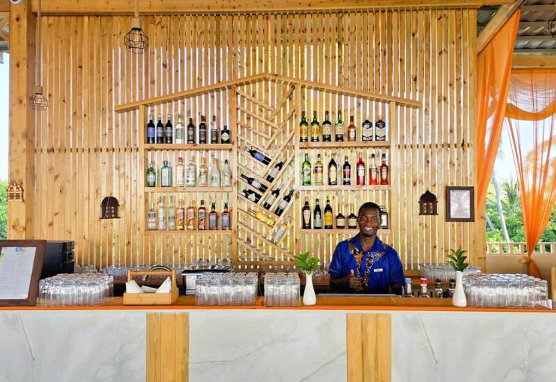 Tropical hotel bar with wooden decor, stocked liquor shelves, orange sheer curtains, and a smiling bartender ready to serve guests