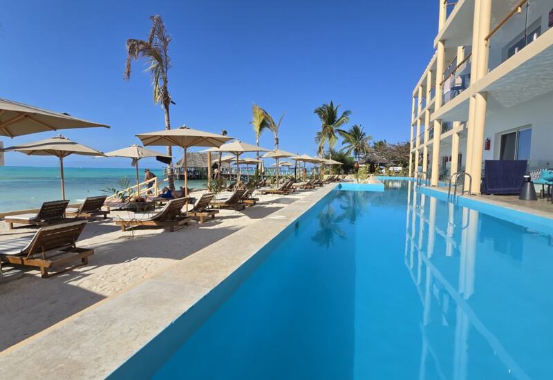 Beachfront pool with lounge chairs and umbrellas on sandy shore, adjacent to a modern hotel with balconies and ocean views