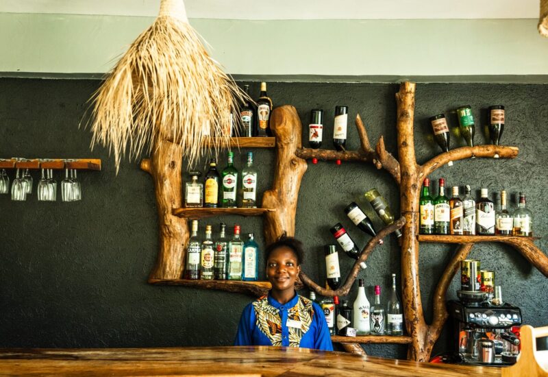 Warm hotel bar with natural wood shelves shaped like a tree, stocked with spirits, and a smiling bartender in vibrant uniform