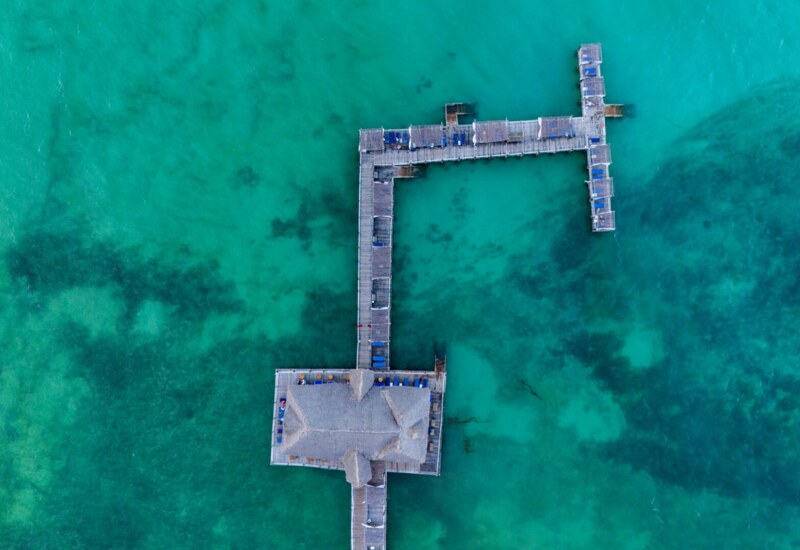 Aerial view of a wooden pier with lounge chairs extending over clear turquoise ocean water, perfect for relaxation and sunbathing