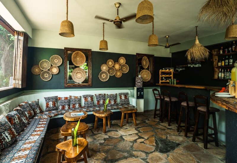 Cozy hotel lounge with patterned cushions on a curved bench, rustic wooden stools, woven basket wall art, and a bar with assorted drinks.
