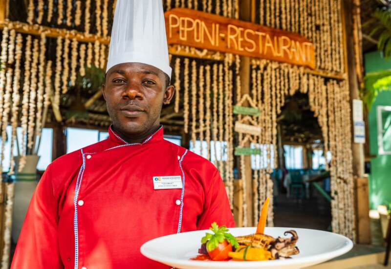 Executive chef in red uniform presenting a gourmet seafood dish at rustic Peponi Restaurant with natural decor and open-air seating