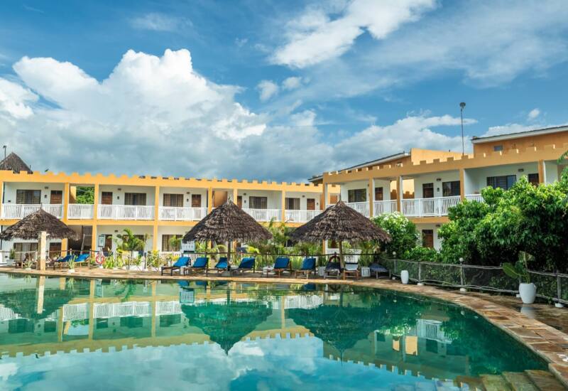 Resort pool area with thatched umbrellas, blue lounge chairs, and two-story yellow guest buildings surrounded by lush greenery