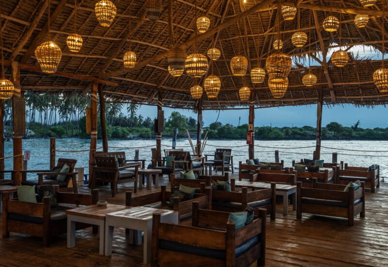 Open-air waterfront lounge with wooden furniture, woven hanging lanterns, thatched roof, and views of palm trees at dusk