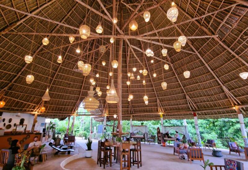 Open-air lounge with high thatched roof, hanging lanterns, wooden furniture, and guests relaxing amidst lush tropical greenery.