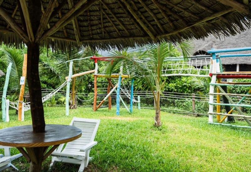 Outdoor seating area with white chairs and wooden table under a thatched roof, overlooking colorful wooden playground in tropical garden