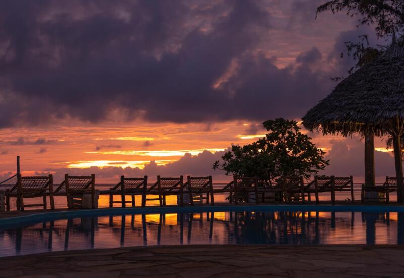 Oceanfront pool area with wooden lounge chairs and thatched umbrellas reflecting a vibrant sunset sky