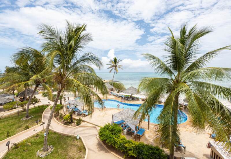 Tropical resort pool area with palm trees, shaded cabanas, lounge chairs, and a beach with calm ocean waters under a partly cloudy sky
