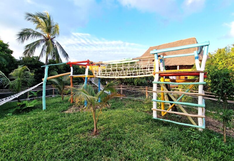 Colorful outdoor wooden playground with rope climbing net set in lush tropical garden with palm trees near thatched roof building